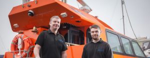Two men standing in front of an orange Goodchild Marine boat.