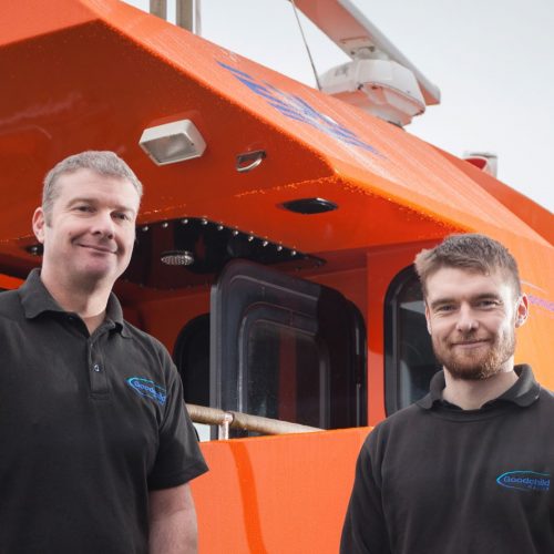 Two men standing in front of an orange Goodchild Marine boat.